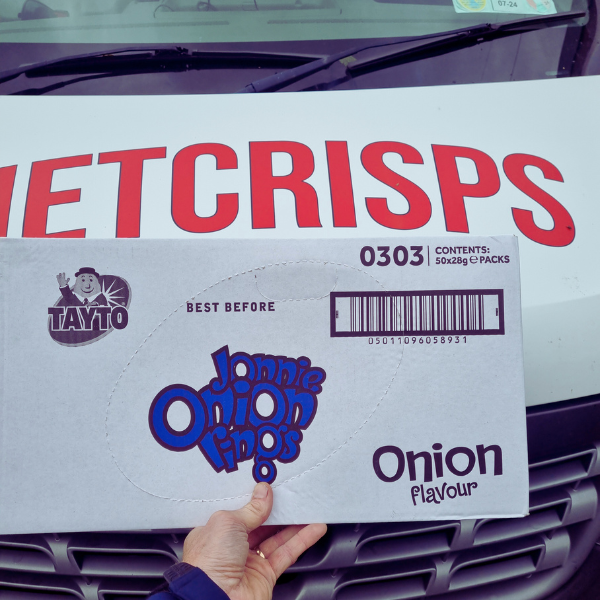 A hand holding a box of Tayto Jonnie Onion rings crisps, featuring onion flavor and a barcode, in front of a delivery van.
