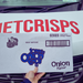 A hand holding a box of Tayto Jonnie Onion rings crisps, featuring onion flavor and a barcode, in front of a delivery van.