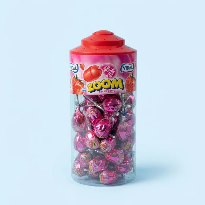 A clear plastic jar of Yirli ZOOM strawberry candy with a red lid, filled with pink and red foil-wrapped lollipops, set against a light blue background.