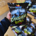 A person holds a bag of Tayto Banshee Bones, a salt and vinegar flavored snack, featuring a spooky design. Other bags are visible in the background.